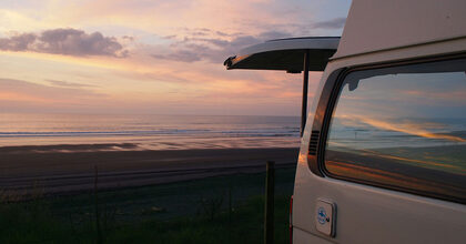 Autocaravana blanca con el portón trasero abierto frente a una playa al atardecer, con el cielo rosado reflejado en la ventanilla