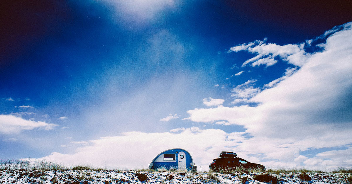 Minicaravana teardrop azul y coche con cofre de techo sobre un campo nevado bajo un cielo azul con nubes