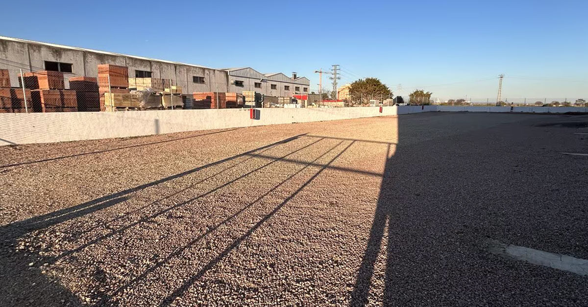 Recinto de parking descubierto con suelo de gravilla y muro perimetral blanco, junto a naves industriales con palés de ladrillos