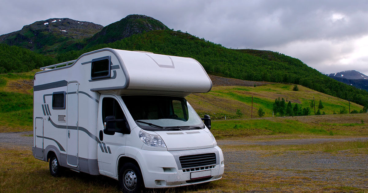 Autocaravana capuchina blanca aparcada sobre hierba frente a montañas verdes con restos de nieve bajo un cielo nublado